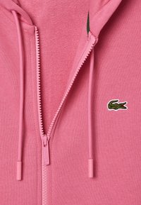 Haut zippé rose avec un tissu texturé, coupe ajustée et un petit logo crocodile brodé en vert sur la poitrine gauche.
