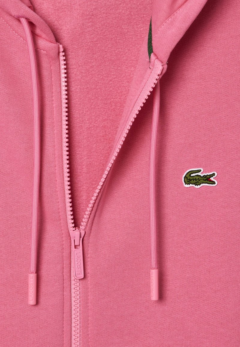 Felpa con zip rosa caratterizzata da un tessuto testurizzato, vestibilità slim e un piccolo logo di un coccodrillo ricamato in verde sul lato sinistro del petto.