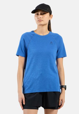 Femme portant un t-shirt de sport bleu à manches courtes, un short noir, une casquette noire et une montre-bracelet, debout devant un fond uni.
