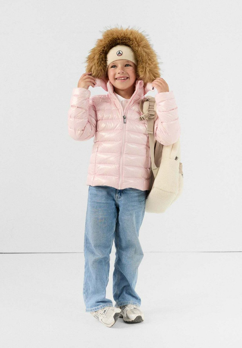 Giacca piumino rosa chiaro con cappuccio foderato in pelliccia, abbinata a jeans in denim azzurro e uno zaino beige. Sneakers bianche completano il look.