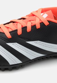 Botín de fútbol negro con una superficie texturizada, cordones de neón naranja y rayas blancas. Presenta tacos moldeados en la suela para mayor tracción.