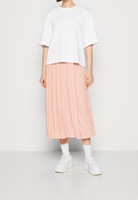 Valkoinen puuvillapaidan t-paita lyhyillä hihoilla, kerroksittain vaaleanpunaisen taitteisen midi-hameen päällä, yhdistettynä valkoisiin lenkkareihin ja nilkkasukkasiin.