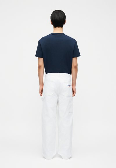 Άτομο με κοντά μαύρα μαλλιά, φορόντας μπλε σκούρο κοντομάνικο μπλουζάκι και λευκό παντελόνι φαρδύ, στέκεται μπροστά σε απλό λευκό φόντο.