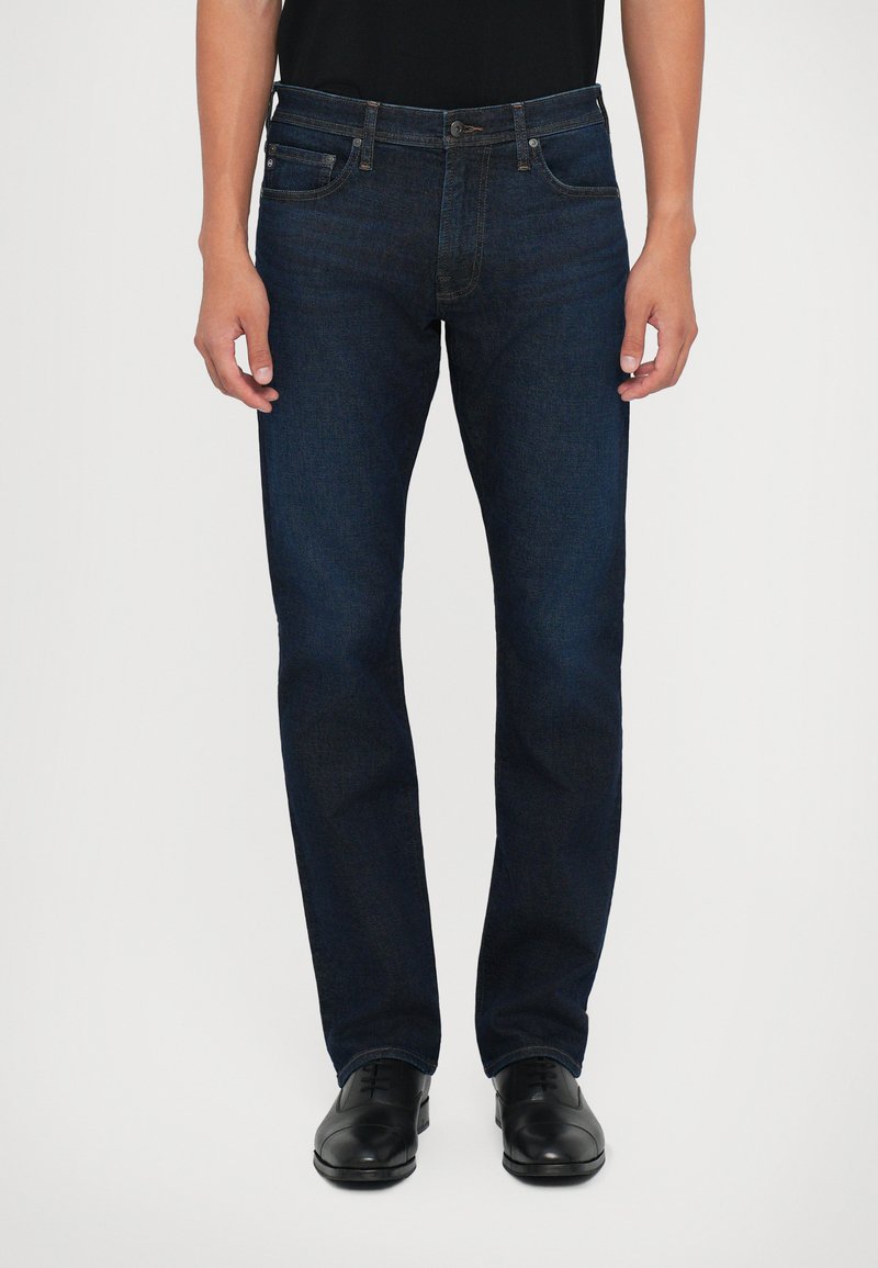 Temno modri jeans z ravnim krojem, dizajnom s petimi žepi in svetlimi šivi. Ujemajo se z črnimi usnjenimi čevlji.