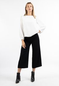 Camicia bianca a maniche lunghe con dettagli arricciati, abbinata a pantaloni neri a gamba larga. La modella indossa stivaletti neri. Sfondo bianco semplice.