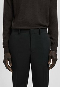 Osoba ubrana w ciemnobrązową koszulę z długim rękawem włożoną do wysokich czarnych eleganckich spodni, stojąca z rękami luźno opuszczonymi wzdłuż ciała.