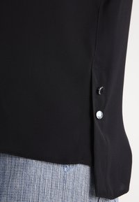 Tessuto di blusa nera con una texture liscia, caratterizzata da un orlo curvo e dettagli con bottoni argentati sui lati.