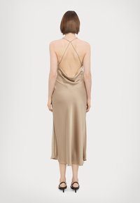Cowl-back midi dress in beige satin, featuring thin straps and a flowing silhouette. Worn with black heeled sandals.
