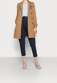 Trench-coat beige, à double boutonnage avec des boutons noirs. Associé à un pantalon court bleu marine foncé et des sandales plates bleu clair.