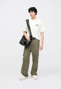 Hvid grafisk t-shirt, olivengrøn cargobukser med lommer, sort skuldertaske og hvide sneakers, med enkelt design og afslappet pasform.