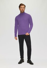 Paarse coltrui van zachte stof, aansluitend ontwerp, gecombineerd met zwarte skinny jeans en zwarte leren laarzen, staande tegen een neutrale achtergrond.