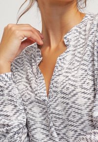 Blouse blanche avec motif en zigzag gris ; présente une coupe ample, un décolleté en V et des poignets froncés. Tissu doux avec un léger éclat.