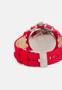 Red textile watch strap with stainless steel hardware, featuring a round watch face and a large case. Back engraved with "DIESEL."