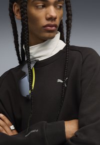 Felpa nera con logo PUMA, colletto oversize bianco e occhiali da sole grigi con dettagli gialli. Anello d'argento alla mano, capelli intrecciati.