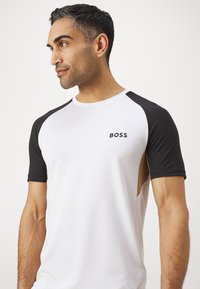 Pánské sportovní tričko v bílé barvě s černými raglánovými rukávy. Na hrudi je logo "BOSS". Hladká textura, moderní design s bočními výřezy.