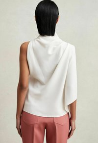 Top alb fără mâneci, cu textură drapată, având un design asimetric. Purtați cu pantaloni roz, adaptați pe corp. Material neted și linii clare.