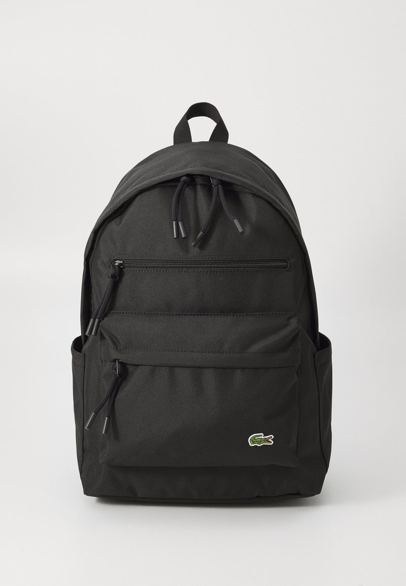 Sac à dos noir en nylon avec un dessus arrondi, deux poches zippées à l'avant et des poches latérales. Comprend des fermetures à cordon et un petit logo à l'avant.