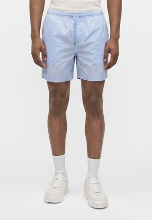 Homme portant un short bleu clair à cordon, des chaussettes blanches montantes et des baskets blanches, debout devant un fond blanc uni.