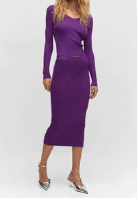 Femme portant un haut ajusté à manches longues violet et une jupe midi côtelée assortie avec des escarpins argentés à bout pointu et bride de cheville, se tenant devant un fond blanc.