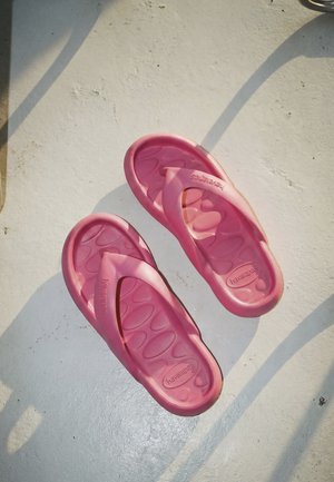 Egy pár kopott rózsaszínű Havaianas papucs fehér, texturált felületen, melyen árnyékok vetődnek átlósan.