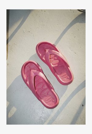 Egy pár kopott rózsaszínű Havaianas papucs fehér, texturált felületen, melyen árnyékok vetődnek átlósan.