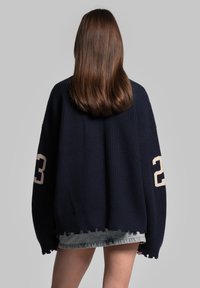 Maglione a coste blu navy con vestibilità oversize, caratterizzato da orlo sfrangiato e il numero bianco "23" sulla manica sinistra. Indossato sopra pantaloni shorts in denim strappati.