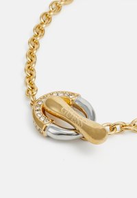 Goldene Kettenhalskette mit einem Toggle-Verschluss, verziert mit Kristallakzenten und einem silberfarbenen Schlaufen-Element. Mit eingraviertem "GUESS"-Detail am Verschluss.