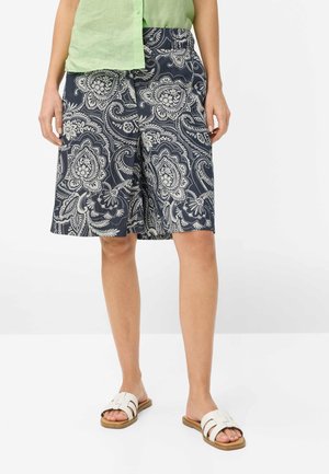 Femme portant un short bleu marine mi-long avec imprimé paisley floral blanc, un haut sans manches vert clair et des sandales blanches à enfiler.
