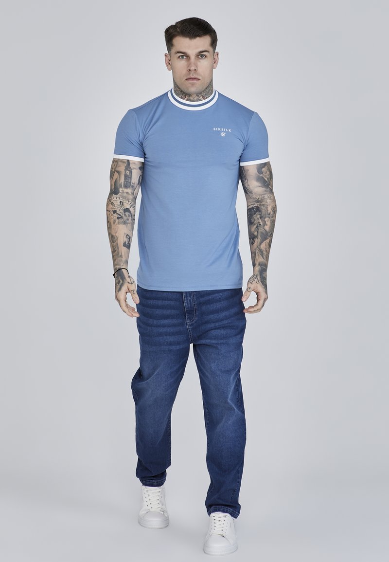 Camiseta de manga corta azul claro con cuello y mangas blancas, combinada con jeans de mezclilla azul oscuro y zapatillas blancas.