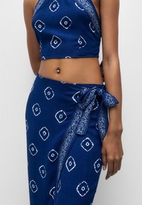 Sininen kaksiosainen kangassetti, johon kuuluuolkaimeton crop-top ja kietaisuhame, joissa on valkoisia geometrisia kuvioita ja sivusiteet.