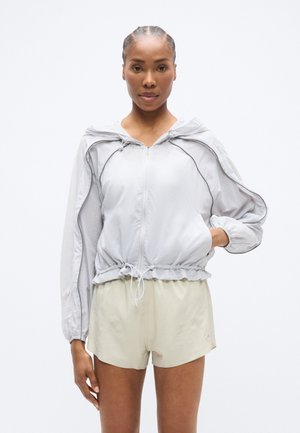 Lys grå lettvektsjakke med hette, innsvingt midje og rynkede ermer; kombinert med beige shorts med detaljer i netting.