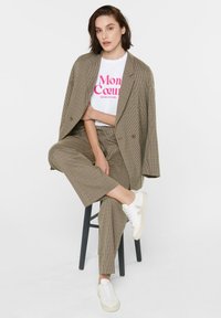 Brown checkered suit with wide lapels, styled over a white graphic t-shirt. Paired with white sneakers. Seated on a black stool.
