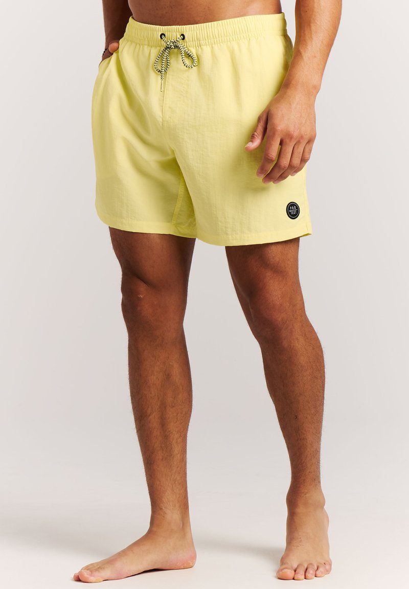 Uomo che indossa pantaloncini da bagno gialli con coulisse e patch con logo, in piedi a piedi nudi contro uno sfondo semplice.