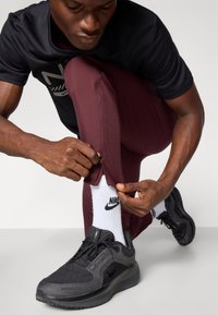Melna sporta T-krekls, bordo krāsas pieguļošas bikses ar rāvējslēdzējiem pie potītēm, melni Nike apavi un balti zeķes ar logotipu, demonstrējot aktīvās apģērba detaļas.