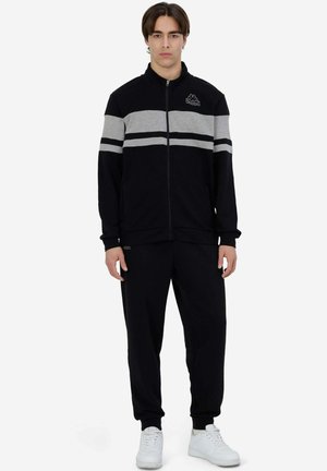 Tuta nera con giacca con zip caratterizzata da strisce orizzontali grigie, tasche laterali e joggers abbinati. Indossata con sneakers bianche.