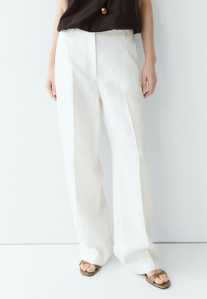 Personne portant un pantalon blanc à jambes larges, un haut marron, un collier pendentif et des sandales ouvertes, debout sur un fond clair.