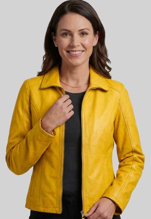 Frau mit braunen Haaren, die lächelt und eine gelbe Lederjacke über einem schwarzen Top vor einem hellgrauen Hintergrund trägt.