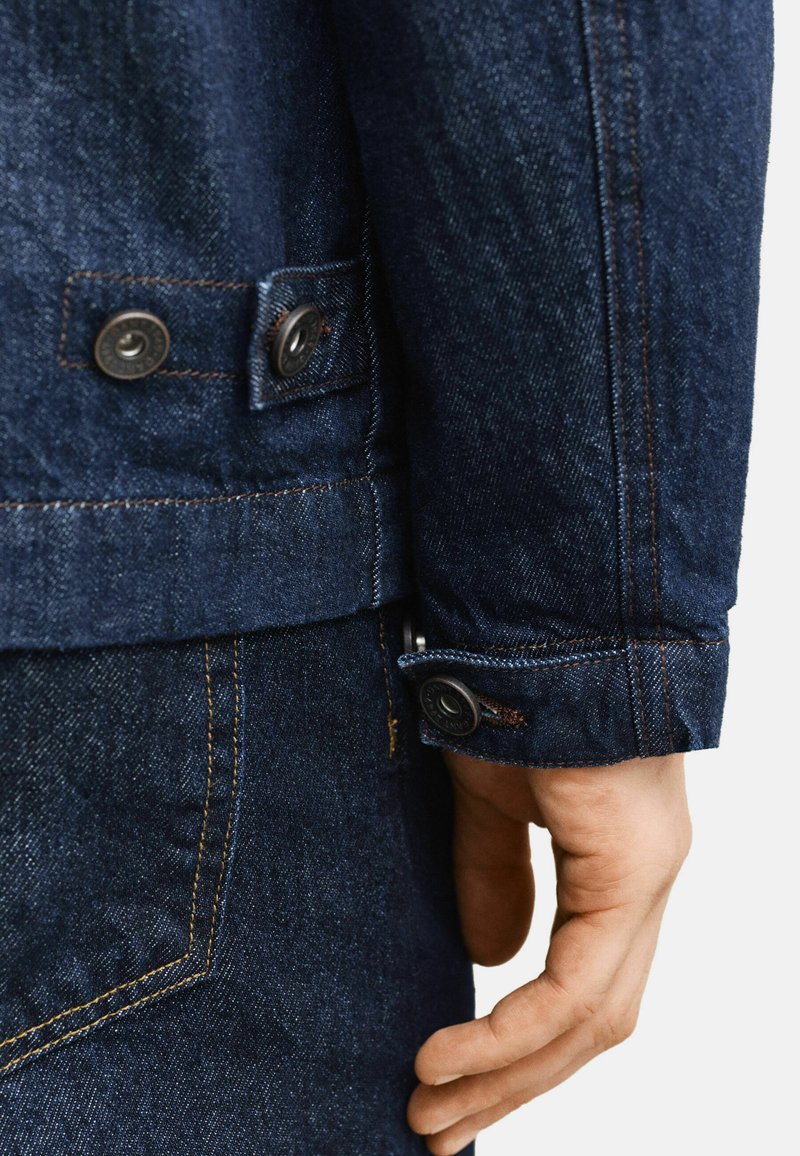 Primo piano della mano di una persona accanto alla manica di una giacca di denim blu scuro con polsino abbottonato e tasca dei jeans in denim coordinato.