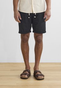 Zwarte shorts met opgerolde manchetten, een tailleband met trekkoord, gecombineerd met brown sandalen met twee gespen en kurk zolen. Achtergrond van houten vloer.