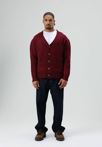Cardigan tricoté bordeaux avec col en V, boutons sur le devant et deux poches ; assorti à un pantalon en jean foncé et des bottes marron.