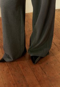 Pantaloni grigi su misura con un design fluido, abbinati a décolleté nere a punta. Il tessuto ha una texture liscia e la vestibilità è a gamba larga.
