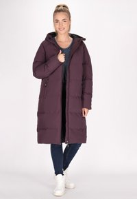 Lungo piumino di colore viola scuro, con cappuccio, zip frontale e tasche laterali. Indossato sopra una maglietta a righe, abbinato a jeans scuri e sneakers bianche.