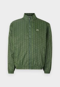 Grüne Zip-Jacke mit geometrischem Muster, hohem Kragen, elastischen Bündchen und einem Lacoste-Logo auf der Brust. Leichtes Material.