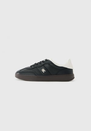 Black low-top sneaker with white padded collar, black laces, brown sole, and a small metallic logo on the side panel.