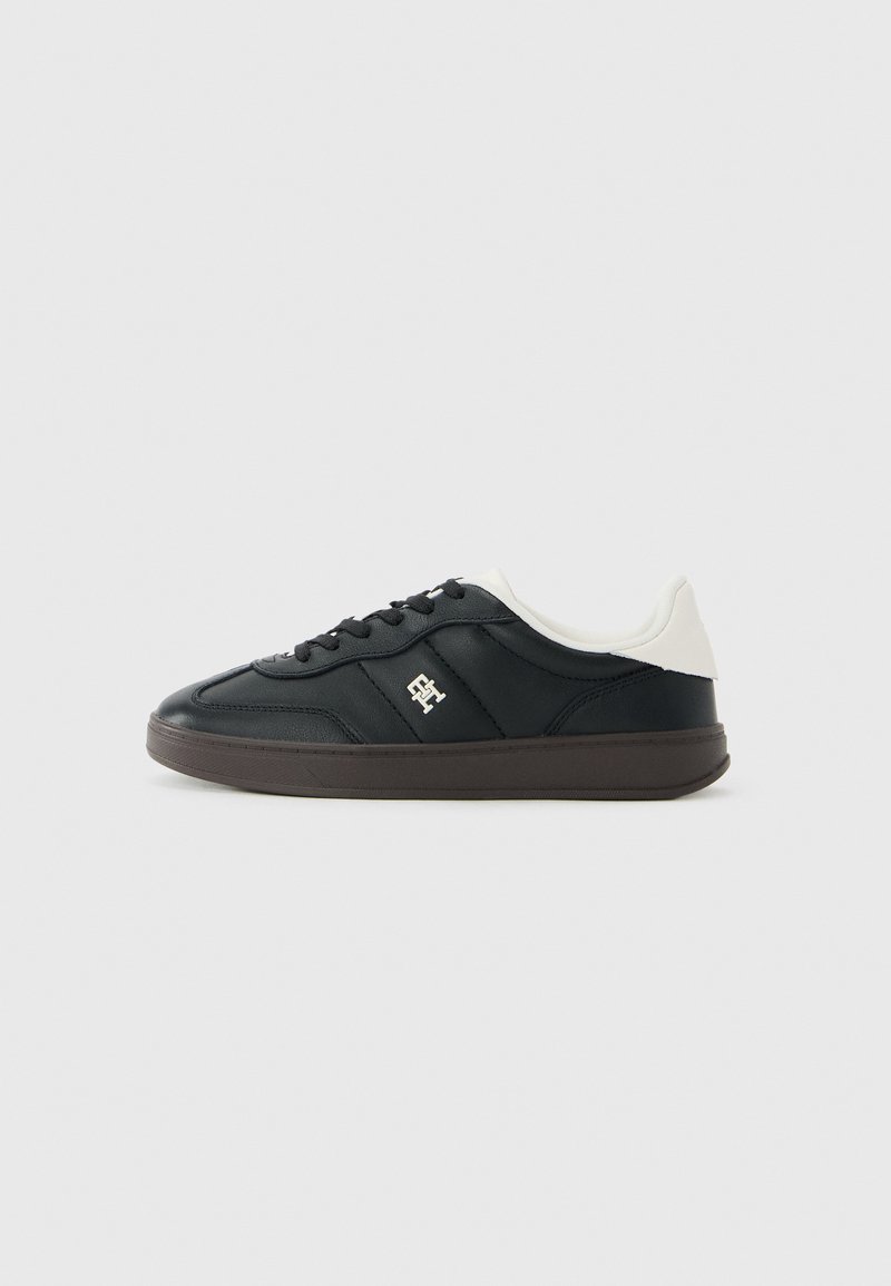 Zwarte lage sneakers met een witte gepluche kraag, zwarte veters, een bruine zool en een klein metalen logo op het zijpaneel.