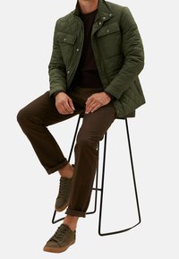 Veste matelassée verte avec deux poches poitrine, chemise marron en-dessous, chinos marron et baskets olive, assis sur un tabouret en métal.