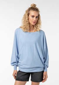 Hellblauer, übergroßer Pullover mit Bootsausschnitt, langen Ärmeln und gerippten Bündchen. Weicher Stoff mit fließendem Fall. Kombiniert mit schwarzen Shorts.