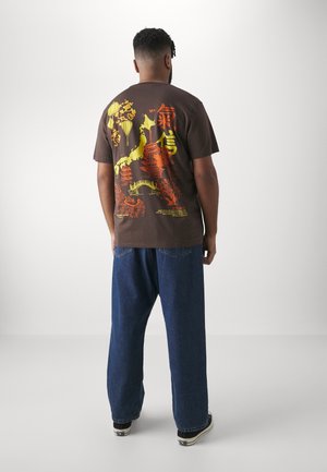Man facing away wearing brown T-shirt with colorful Asian-inspired design and blue jeans against plain white background.