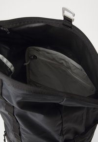 Schwarze Stofftasche mit weit öffnendem Hauptfach. Das Innere verfügt über ein graues gepolstertes Fach mit Reißverschluss. Strukturierte Außenseite mit verstellbaren Trägern.