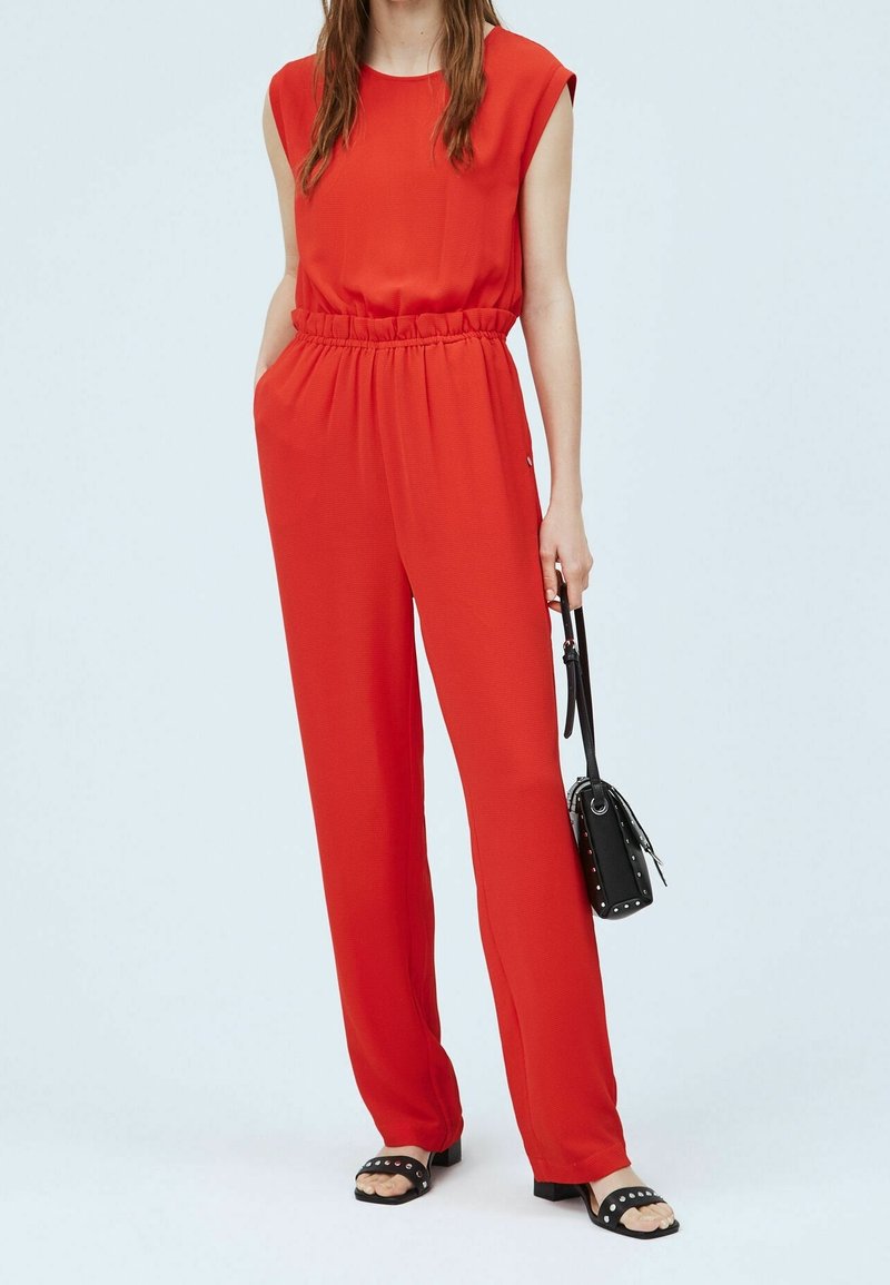 Combinaison rouge à manches courtes, taille élastique cintrée et jambes larges. Associée à des sandales noires cloutées et un petit sac à main noir.